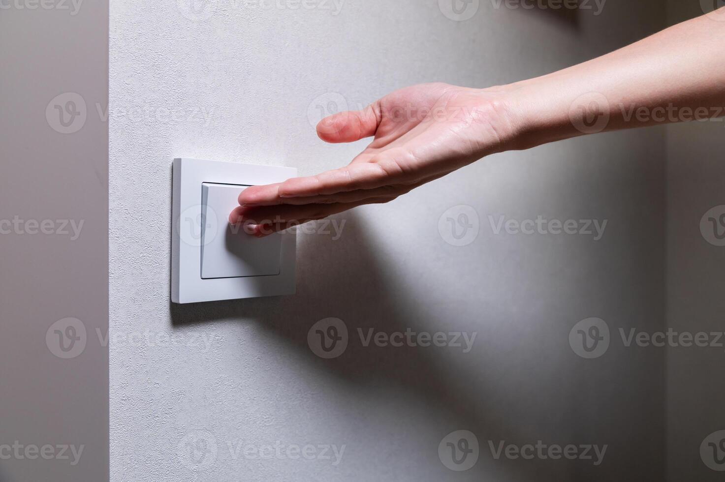 Close-up of an unrecognizable young Caucasian woman's hand turning on a light in a dark room by pressing a switch on a white wall. Close-up photo