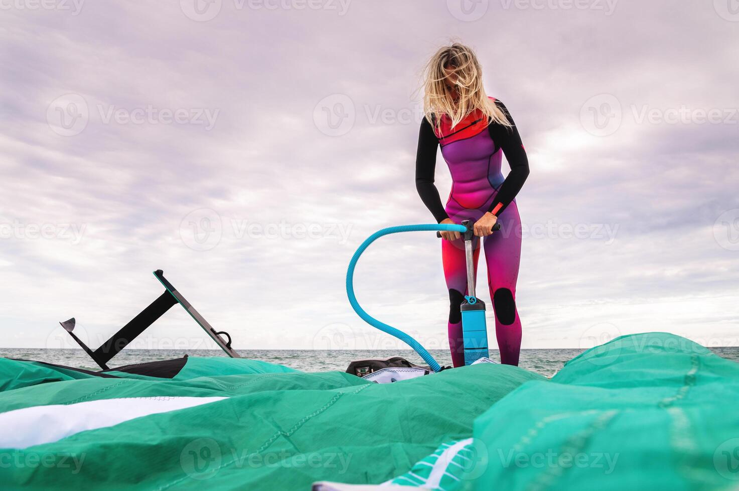 kitesurfer en un traje de neopreno infla su cometa con aire utilizando un bomba en contra el antecedentes de un nublado nublado cielo foto