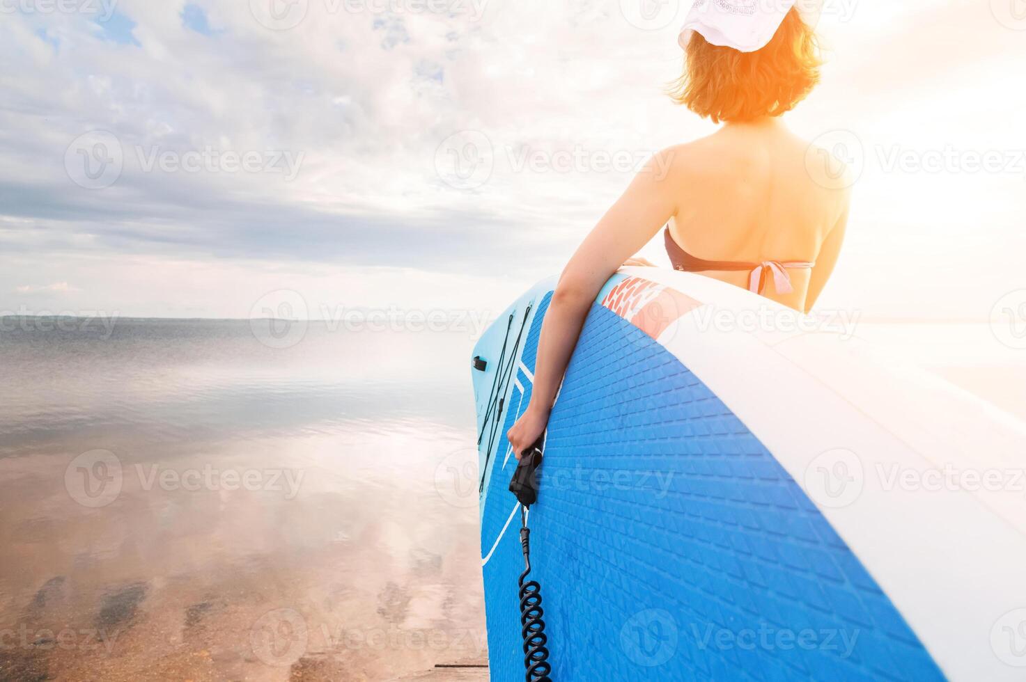 Low angle wide angle young Caucasian woman carries her paddleboard towards the water at sunset on a wooden platform. SUP and surfing concept. photo