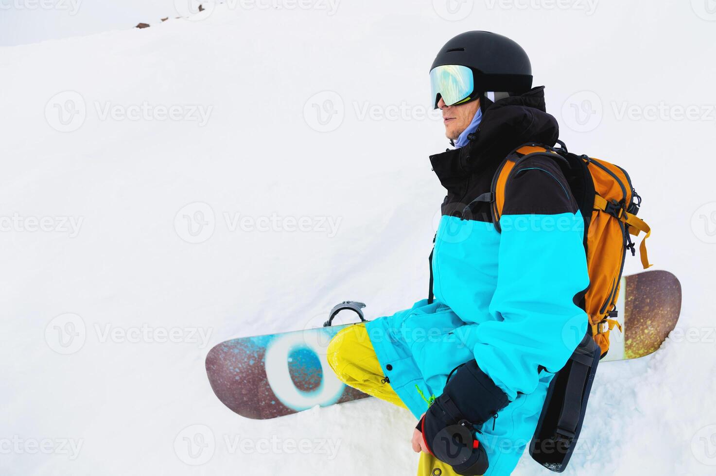 Young man in sportswear and with snowboard sitting on snowy terrain looking towards mountains on cloudy winter day photo