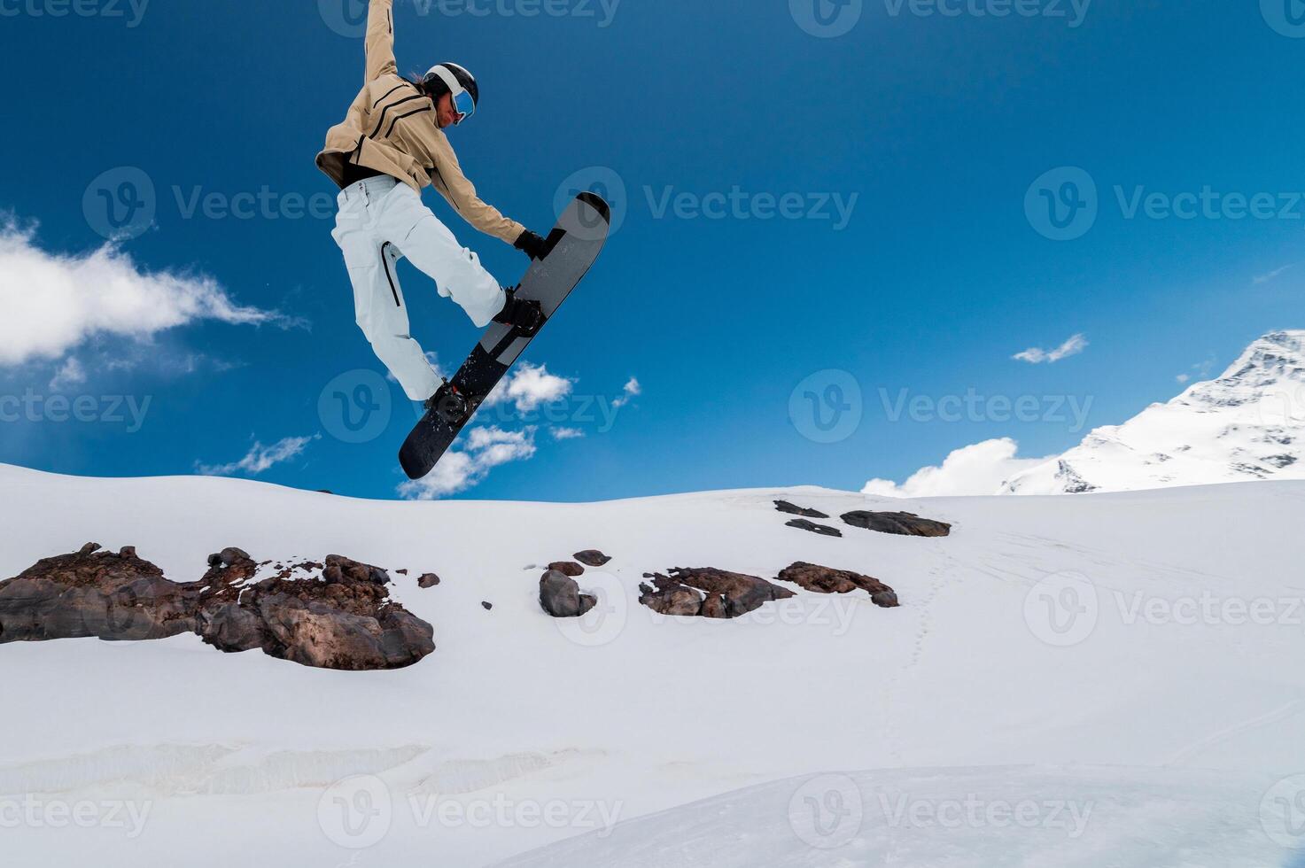 snowboarder jumps into the air against a background of blue sky and mountains. The guy grabs the edge of the board with his front hand while jumping, performing a trick photo
