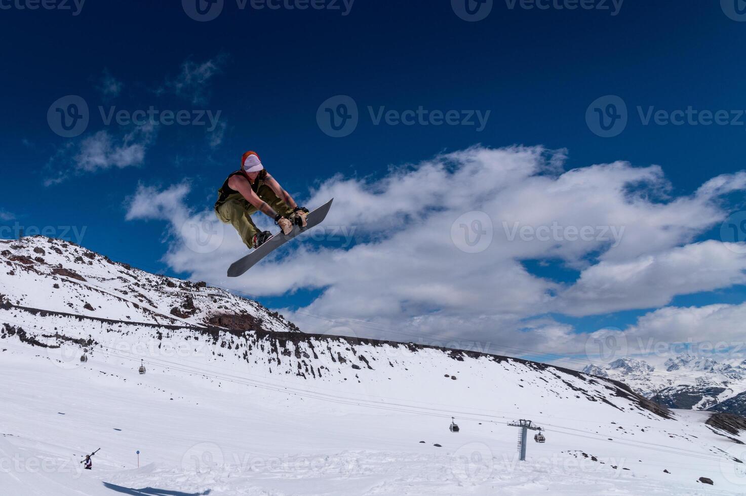 snowboarder jumps from a big air with a board grab, a nose or tail trick in rotation, against the backdrop of snow-capped mountains and the bright sun photo