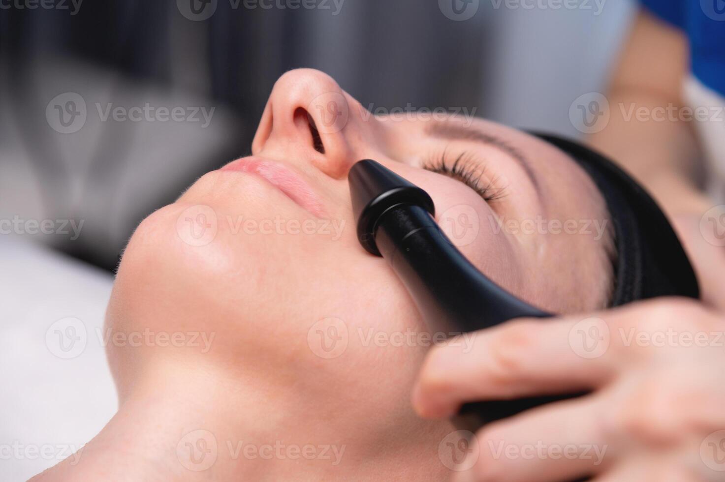 Close up of beautician hand making ultrasound facial procedure by special equipment. Her closed eyes with slight smile photo