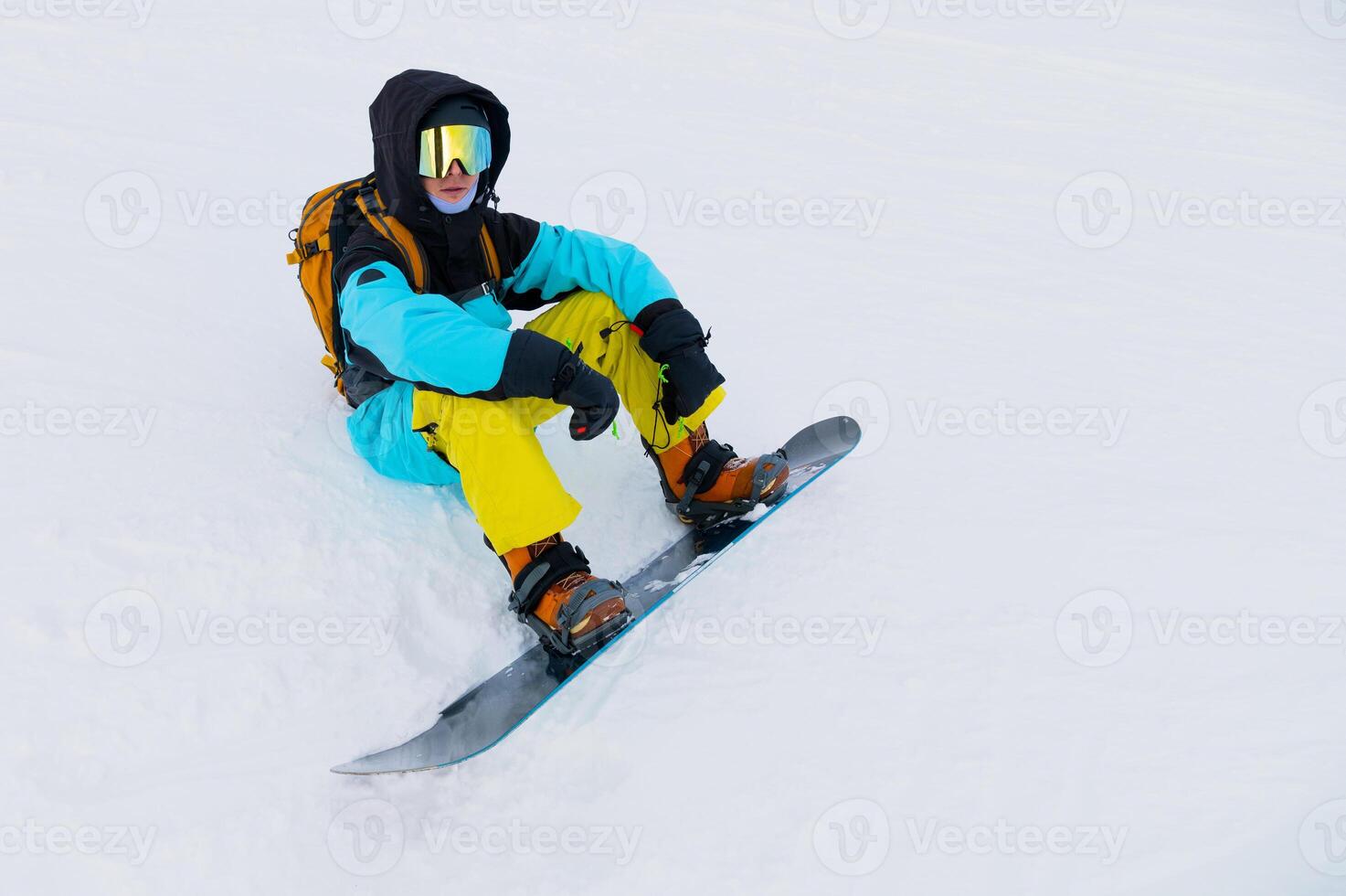 Young man in sportswear and with snowboard sitting on snowy terrain looking towards mountains on cloudy winter day photo
