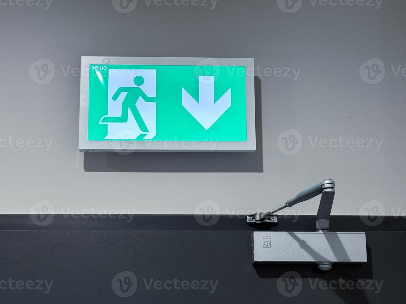 A green emergency exit sign with a figure running and a large arrow pointing down, mounted above a door closer mechanism on a gray wall photo
