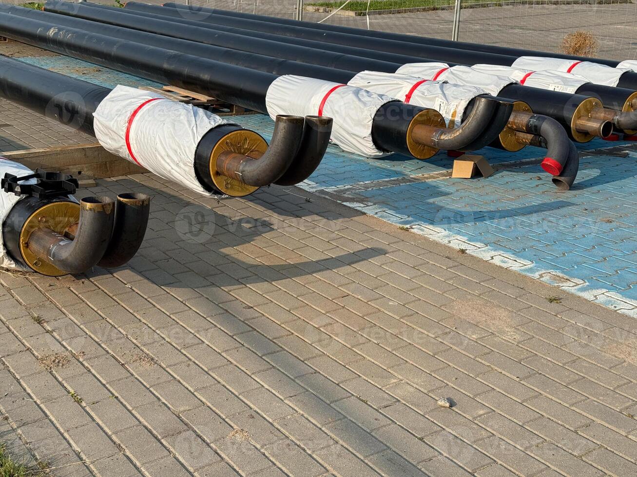 Stacked black pipes with curved ends, partially wrapped in insulation, on a paved construction site photo