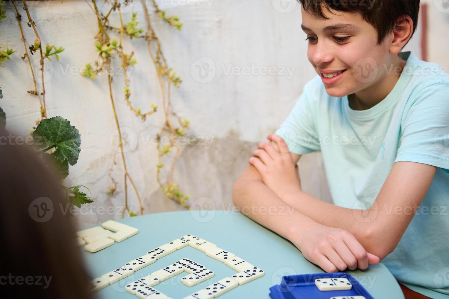 Smiling boy playing dominoes at a table outdoors, enjoying a fun and engaging game with friends or family in a sunny garden setting, showcasing leisure and happiness. photo
