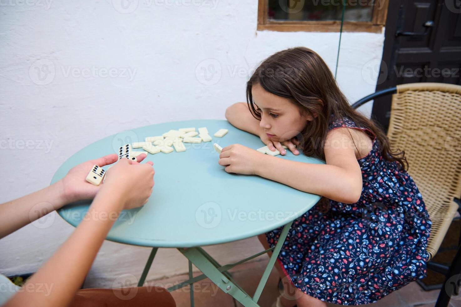 Young girl in summer dress playing dominoes with a family member, reflecting on strategy at a patio table outdoors on a warm day. photo