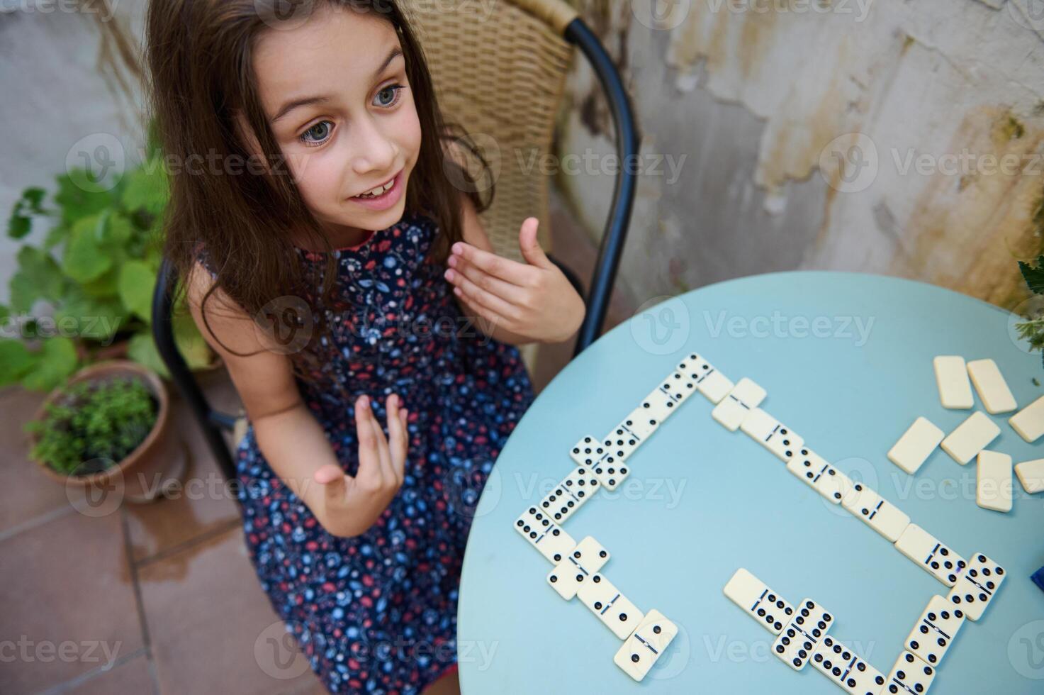 Young girl playing dominoes at a table with excitement and curiosity in an outdoor setting photo
