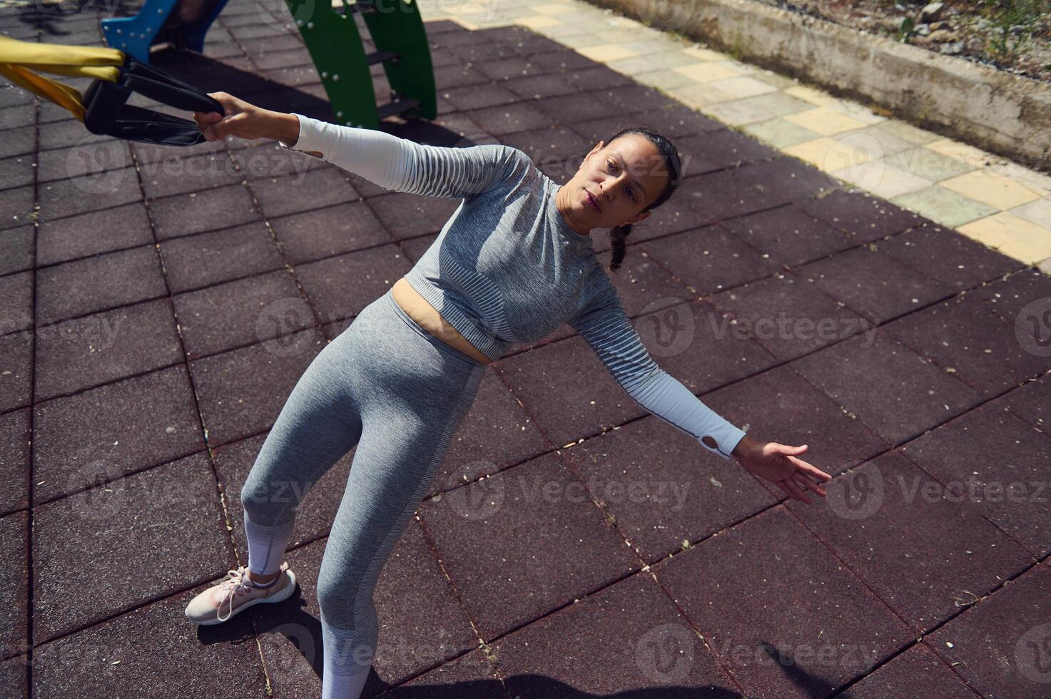 Young woman in athletic wear performing suspension training outdoors on a sunny day, demonstrating strength and balance during a dynamic full-body workout in a fitness park. photo