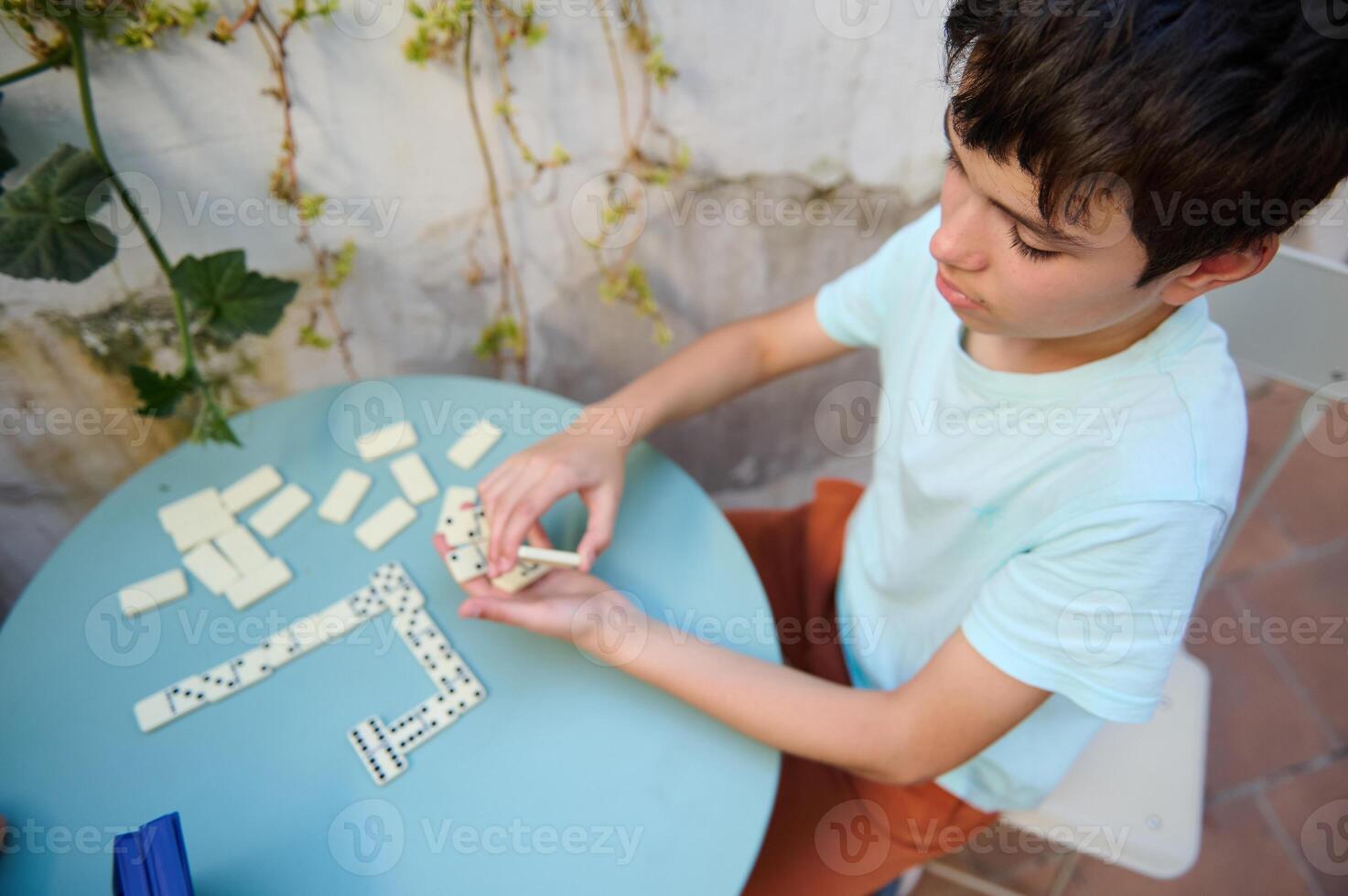 Young boy sitting at a blue table engaged in a game of dominoes outdoors on a sunny day, focusing on strategy and creativity in a serene garden setting. photo