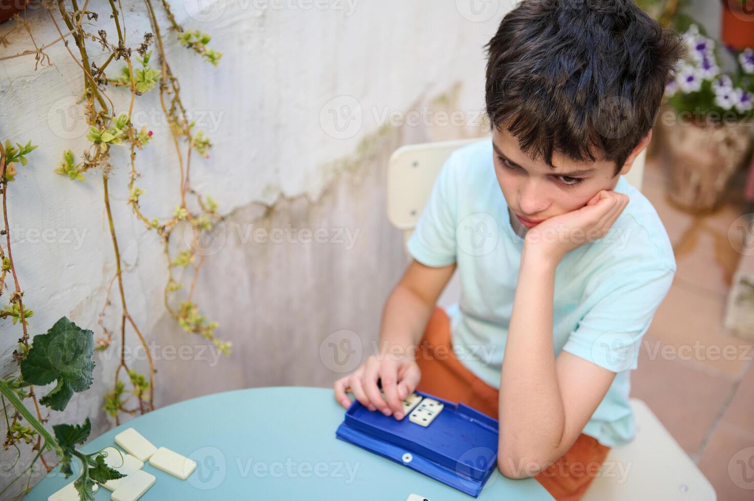 Pensive boy engaged in a thought-provoking game of dominoes at a round table, surrounded by outdoor elements and a hint of greenery, reflecting concentration and contemplation. photo