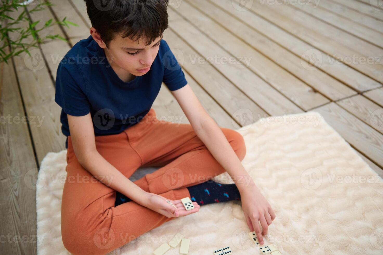 Young boy in a blue shirt and orange pants playing dominoes on a fluffy blanket outdoors, focusing on his next move with concentration and thoughtfulness. photo