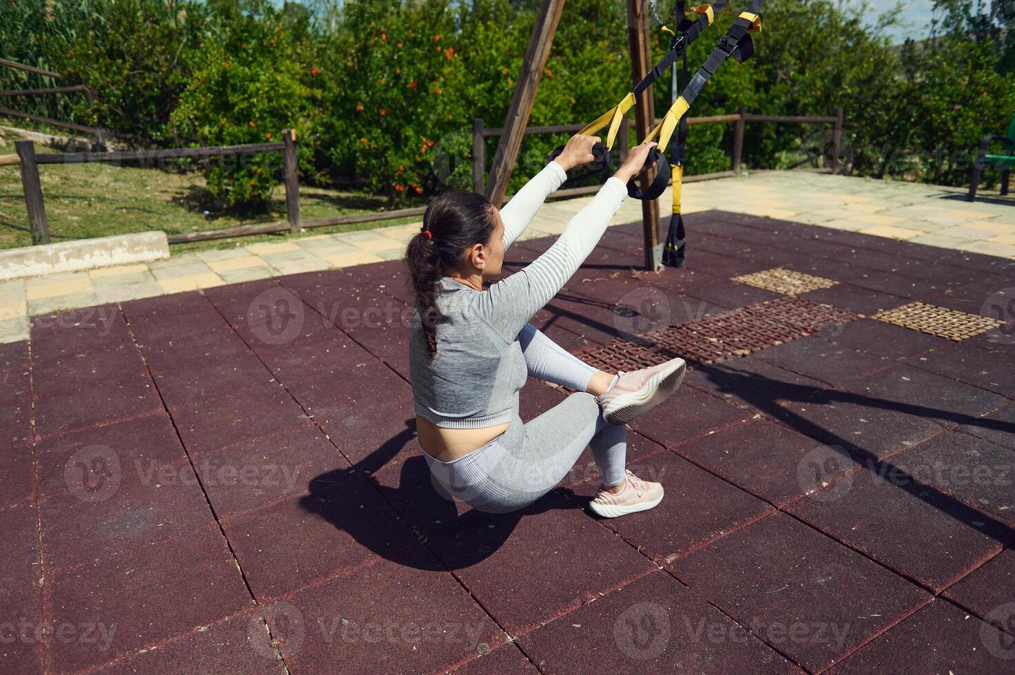 Woman practicing outdoor fitness using suspension trainer in park, performing a balance exercise to enhance strength and core stability photo