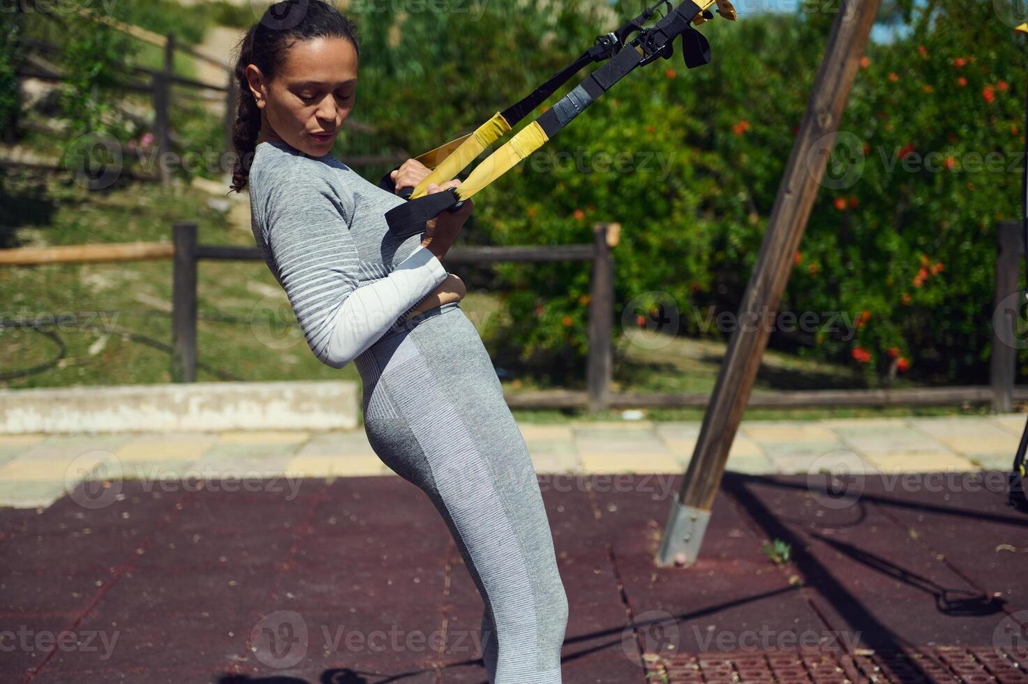 Woman engaging in outdoor TRX workout, focusing on strength and balance in a park setting on a sunny day photo