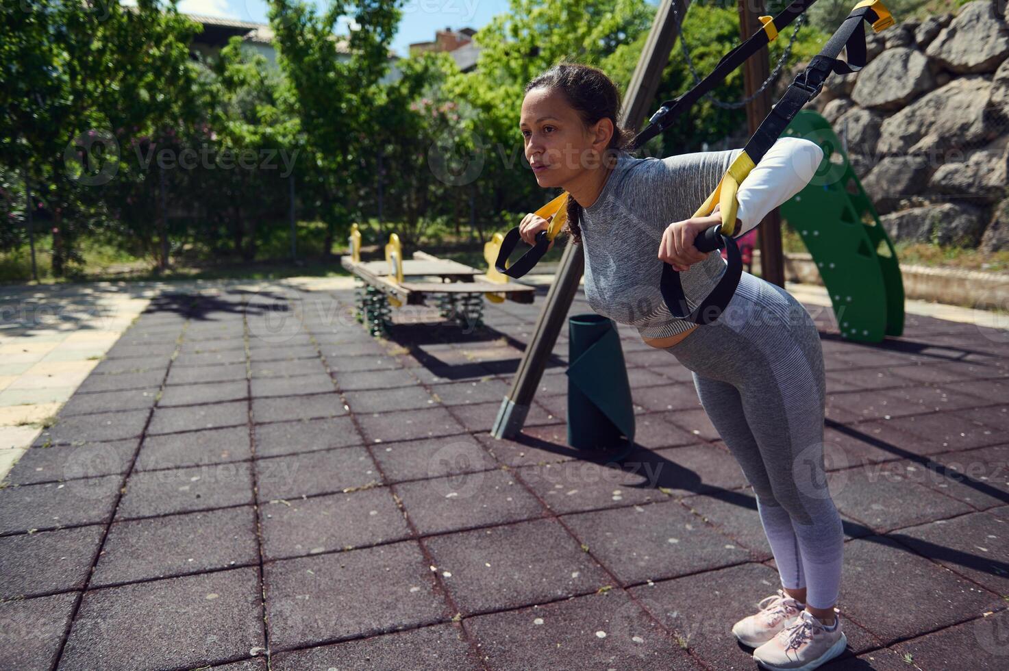Young woman exercising with suspension trainer in outdoor park setting, showcasing strength and determination during workout routine on a sunny day photo