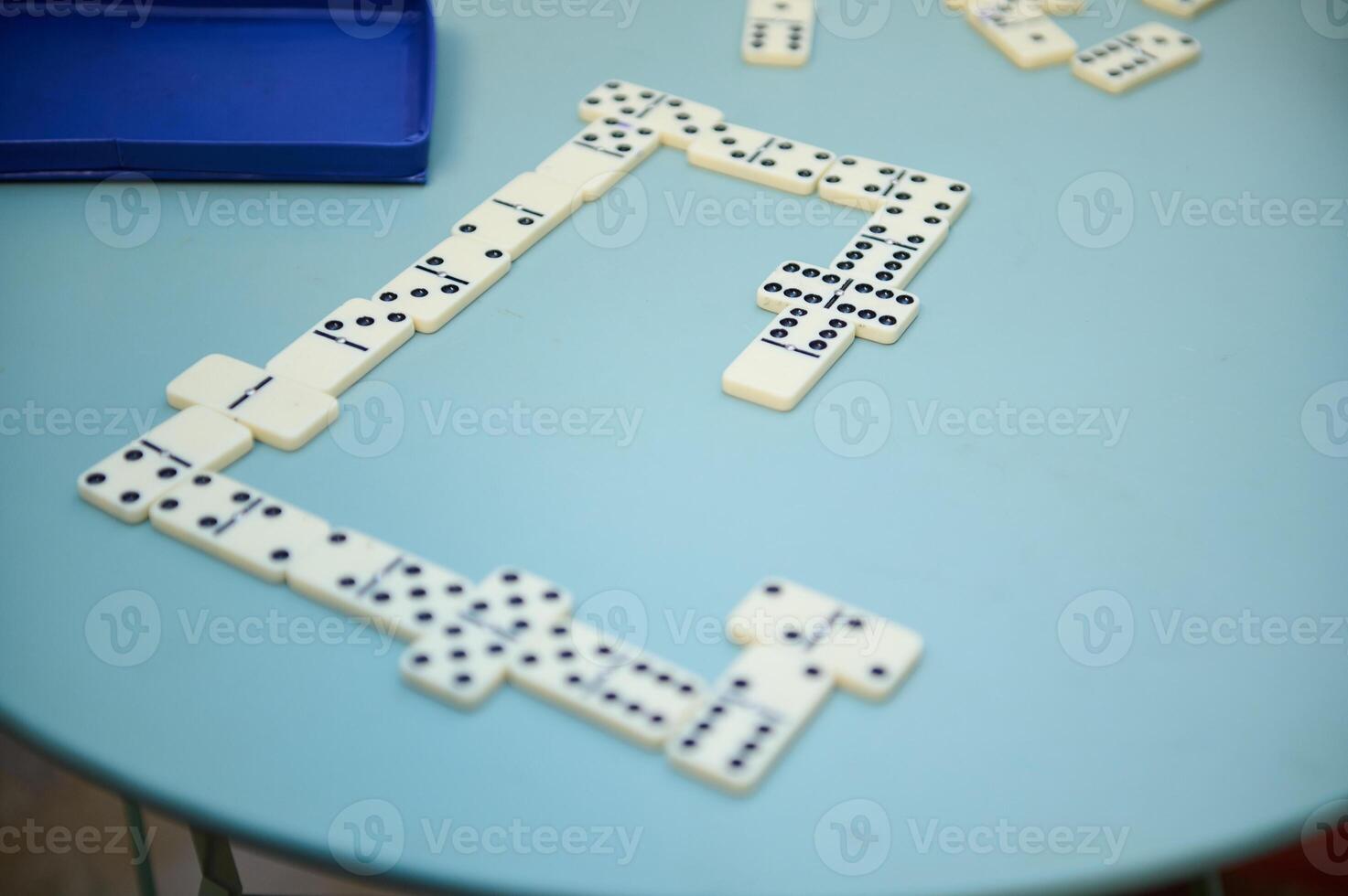 Dominoes game in progress on a round blue table, showcasing a strategic layout with pieces interconnected and box nearby, evoking a sense of leisure and strategy. photo