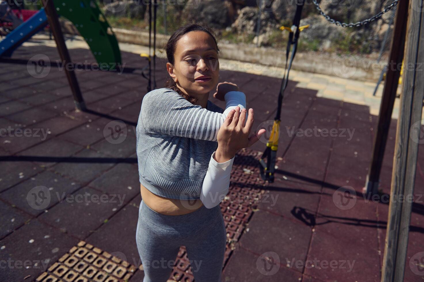 Woman in athletic attire stretching her arm outdoors in a park setting, enjoying a sunny day. Focus on fitness and wellness in an outdoor environment. photo