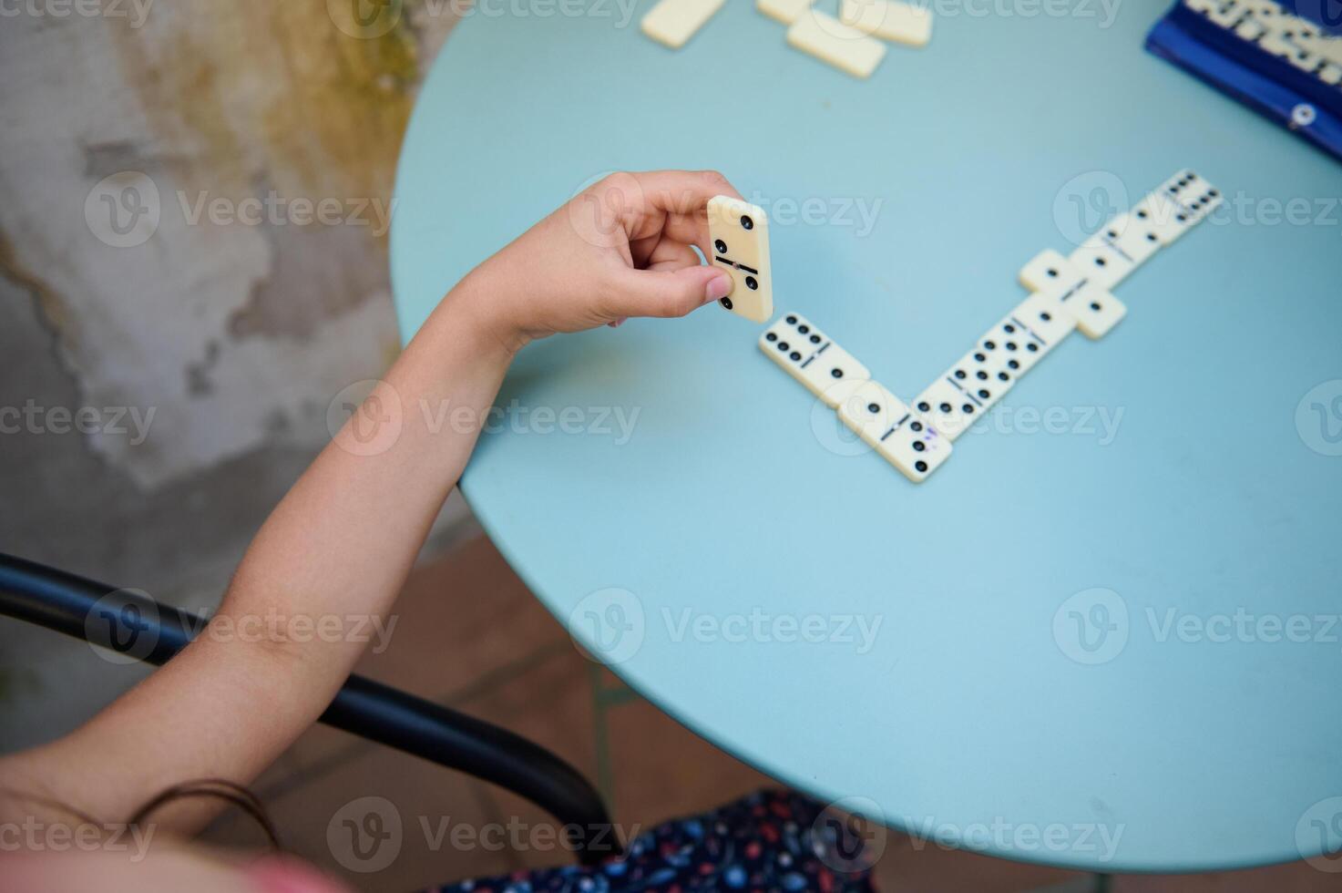 Child playing dominoes at a round blue table outdoors, developing strategic thinking and motor skills while enjoying leisure time in a casual setting on a sunny day. photo