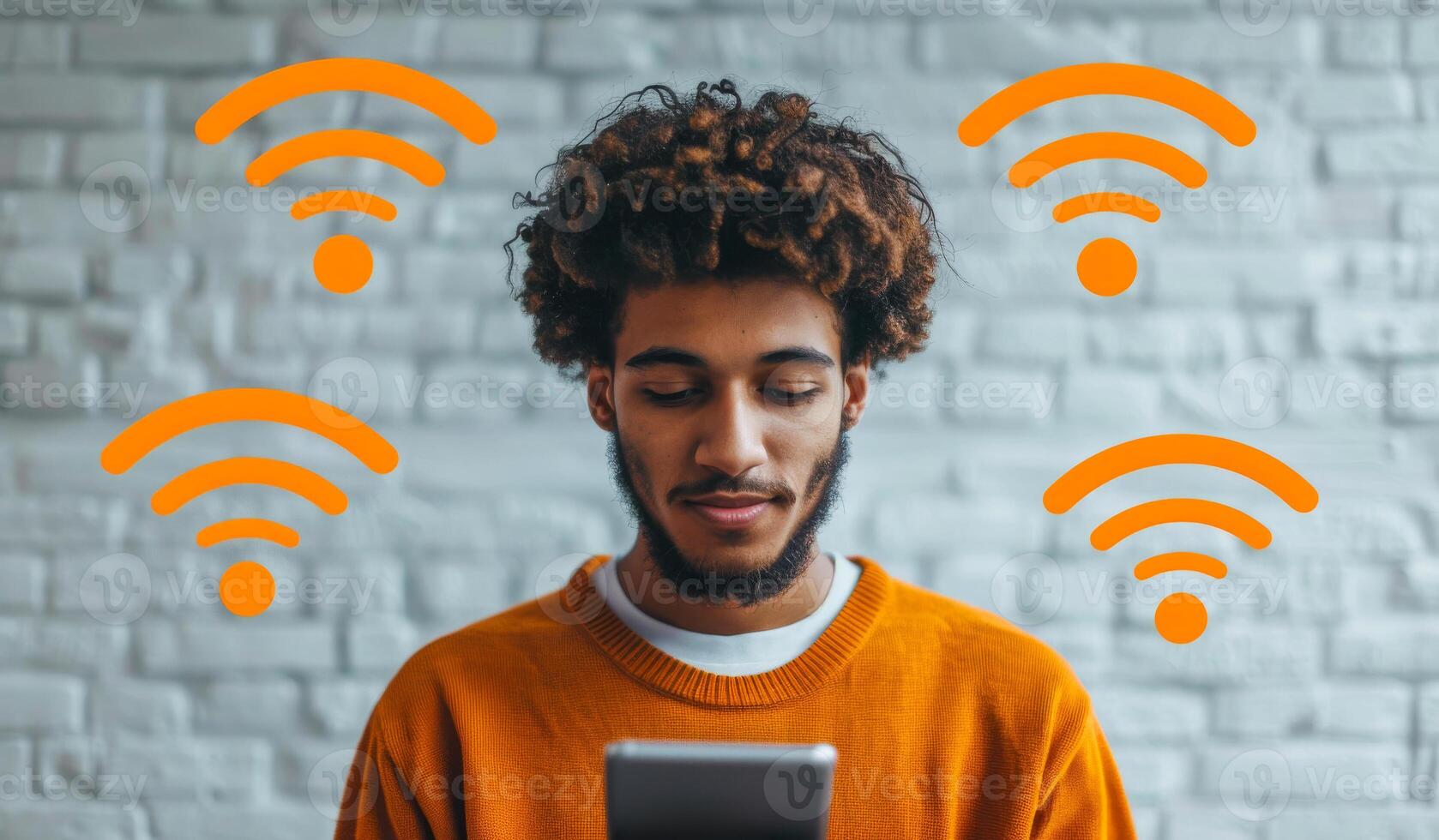 A man is sitting in front of a brick wall with a tablet in his hand. The tablet is displaying a WiFi symbol, indicating that he is connected to the internet photo