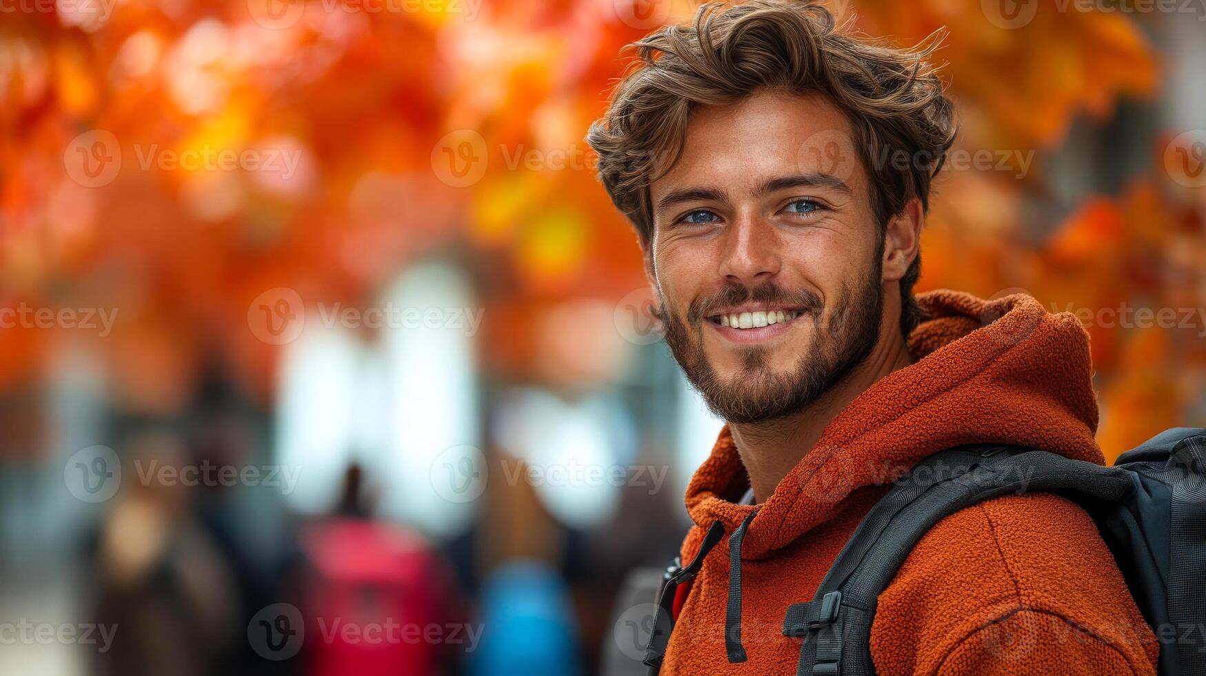 A man with a backpack and a smile on his face. He is wearing an orange hoodie photo