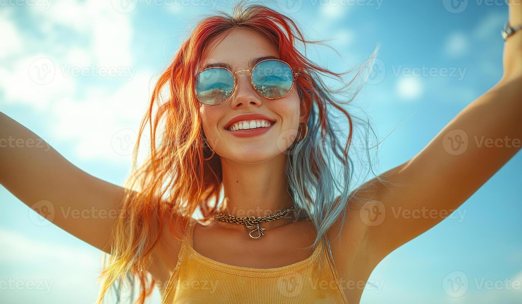 A woman with red hair and blue and green streaks in her hair is smiling and wearing sunglasses. She is wearing a yellow tank top and a necklace photo