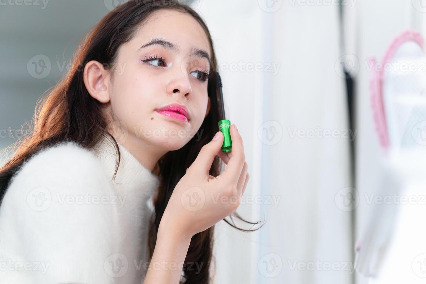 Teenage girl applying makeup, young girl putting some mascara on her eyelashes in front of a mirror photo