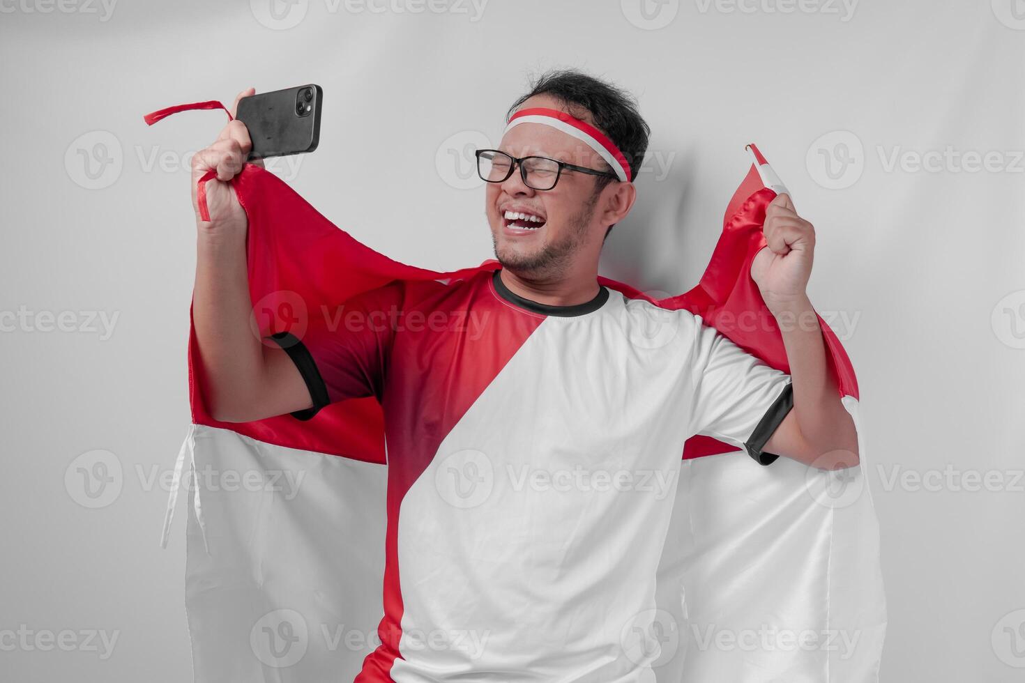 Joyful Indonesian man holding flag over his back and smartphone while raising fist to celebrate. photo