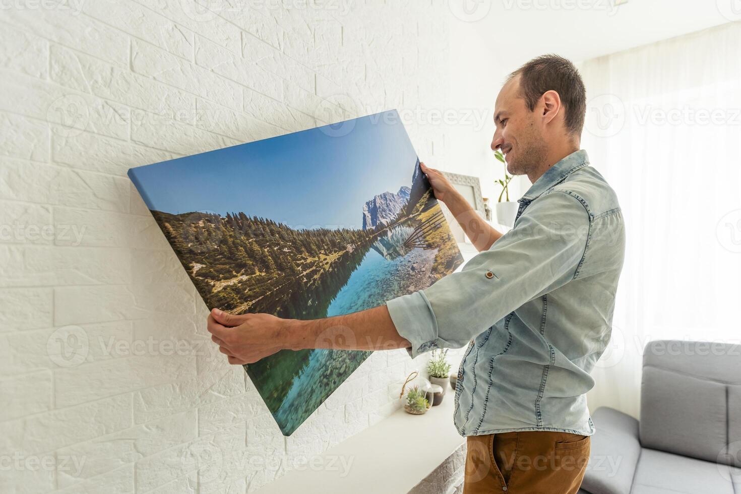 Canvas print with gallery wrap and dry grass interior decor. Woman hangs photography on white wall. Hands holding photo canvas print with image of island in Biarritz city in France