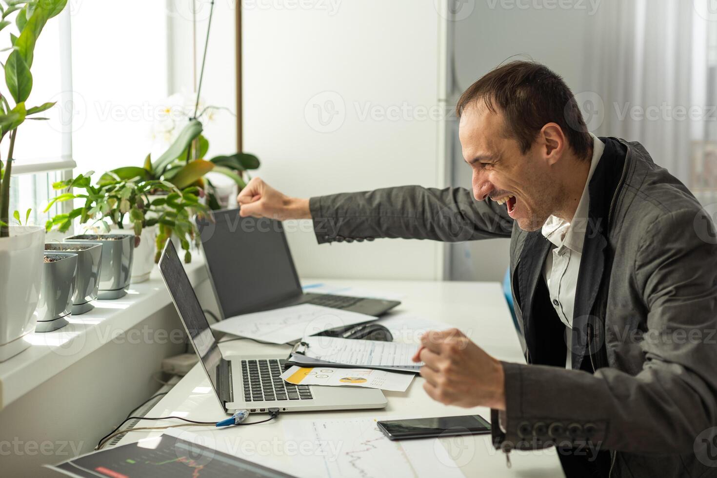 Angry annoyed frustrated caucasian business man, sitting at workplace with laptop, screaming, gesturing with fists, failed deal, disappointed with project result, experiencing negative emotions photo