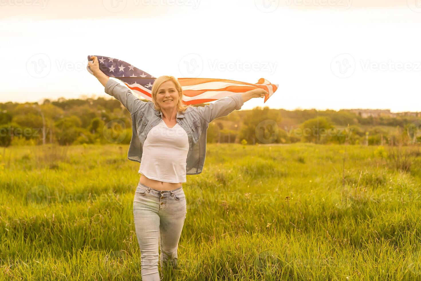 attractive woman holding an American flag in the wind in a field. Summer landscape against the blue sky. Horizontal orientation. photo