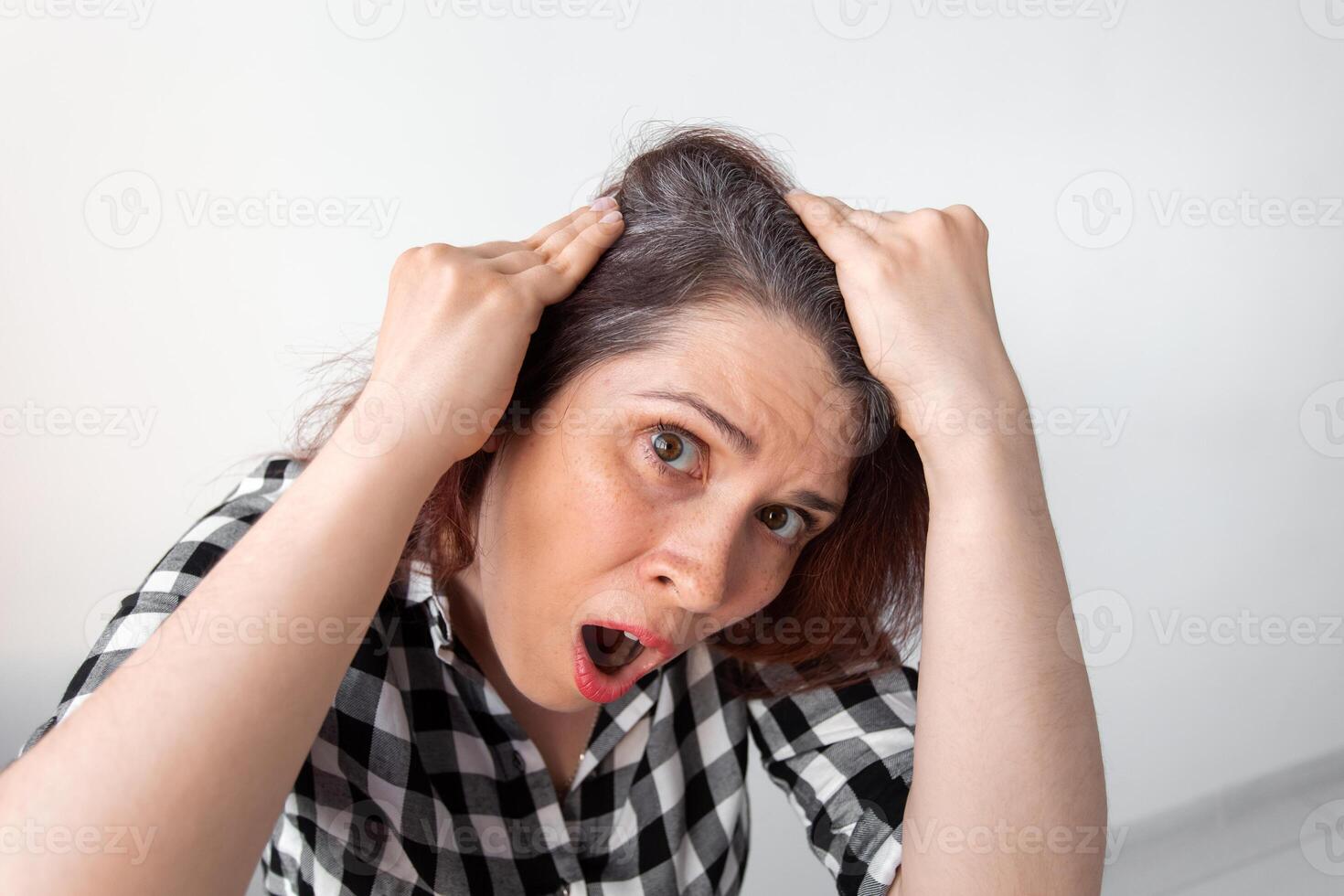 Horrified young woman looking in mirror staring open mouthed at the first grey hair, a first sign of ageing. photo