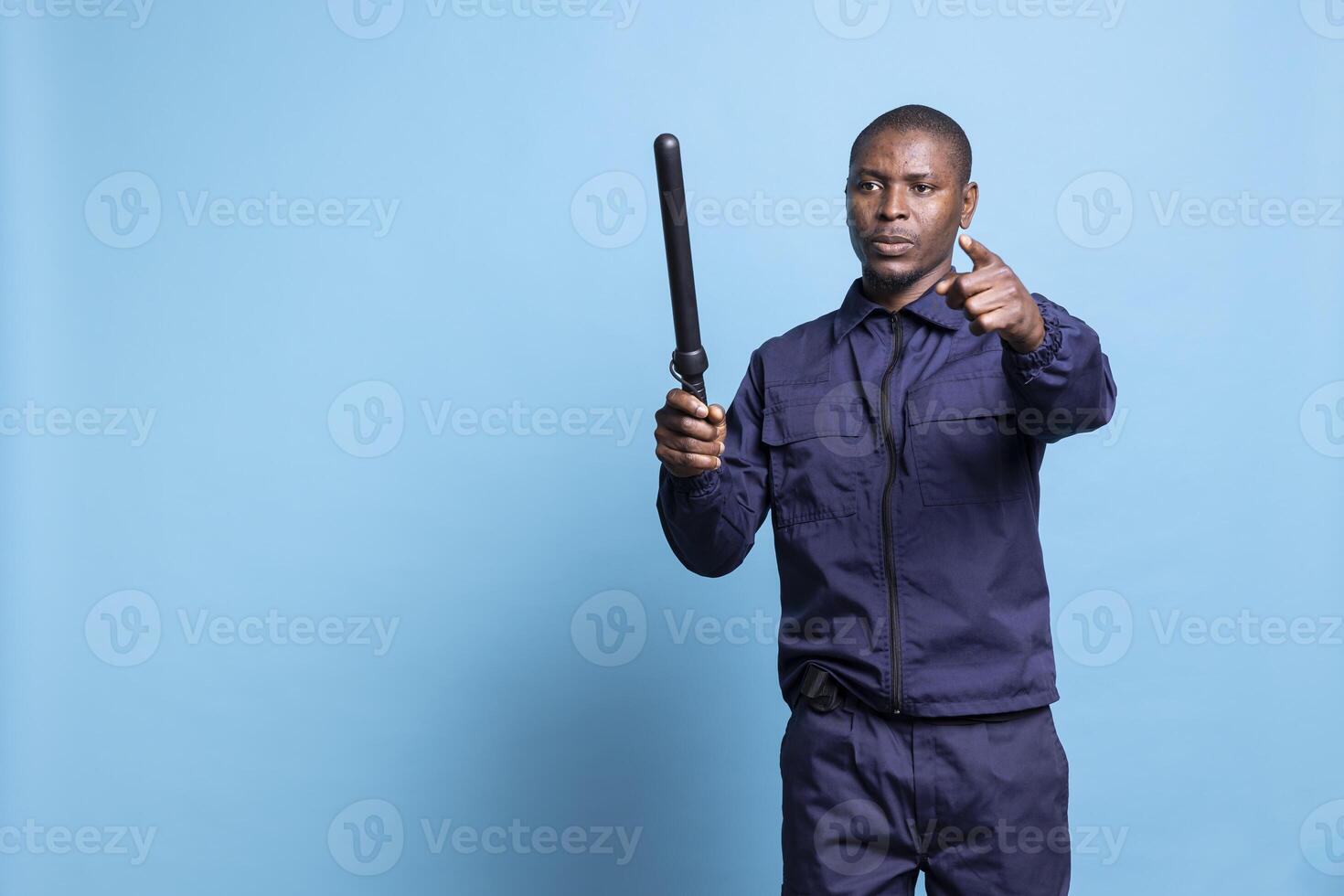 Mall security guard holds a baton pointing at people to approach, standing against blue background. Agent equipped in uniform directing the public, protection duty in studio. photo