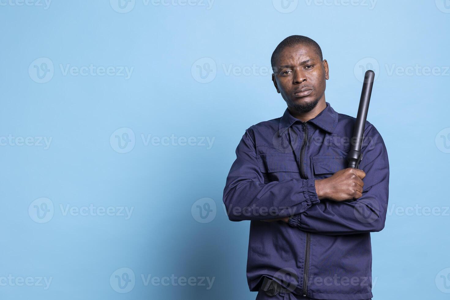 African american security officer poses proudly with his baton on camera, showing confidence with his weapon and professional uniform. Armed bodyguard standing against blue background. photo