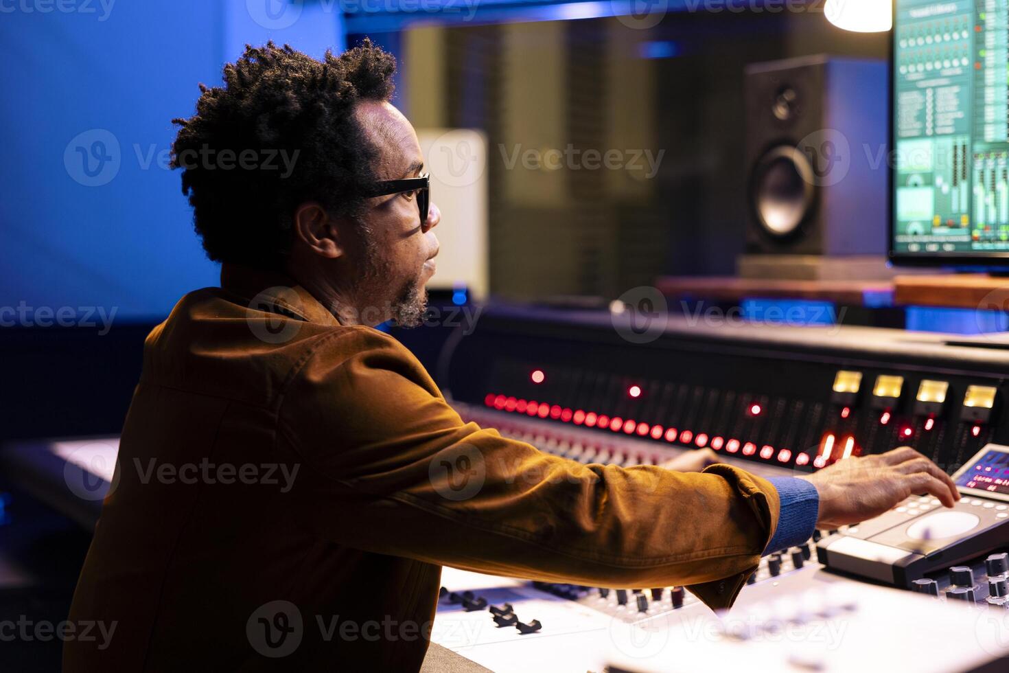 African american record producer adjusting sound settings on control board desk, editing tunes in professional studio. Music engineer working on mixing console to edit music, pushing faders. photo