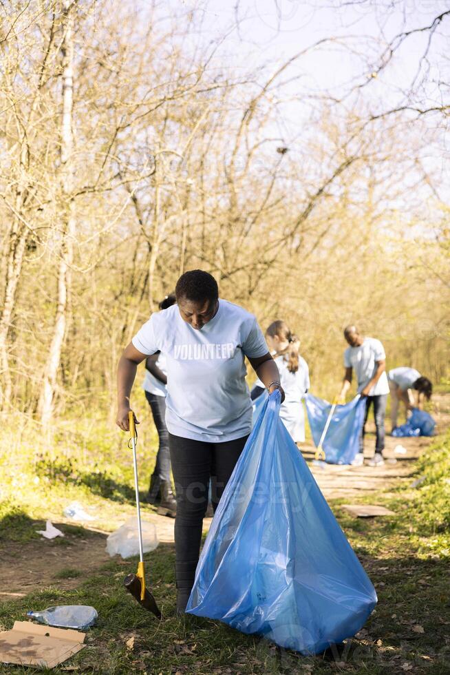 Woman activist grabbing garbage with a claw tool in the forest, clearing habitat of rubbish and plastic junk. African american person doing voluntary work against illegal dumping. photo