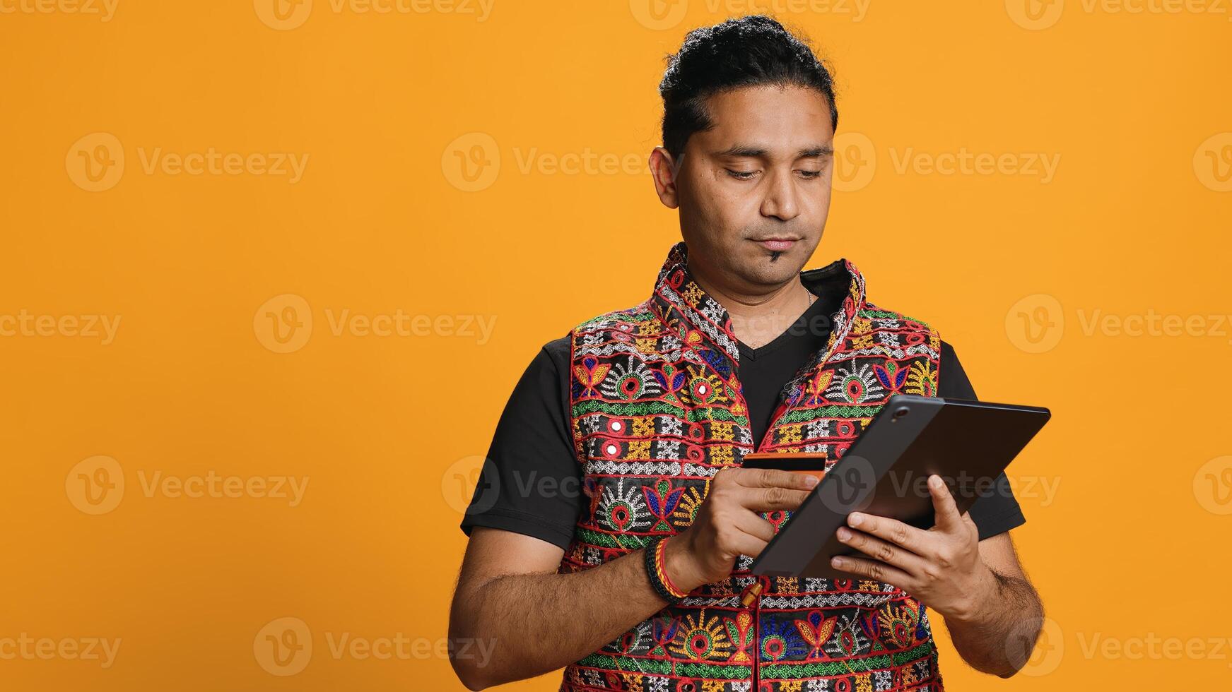 Man adding payment method on shopping website using tablet, taking advantage of promotions, studio background. Indian person buying stuff online using credit card, typing data on device, camera A photo