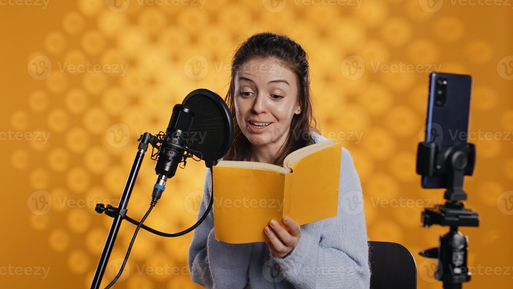 Media star filming vlog at work, reading book to record audiobook, studio background. Voice actor doing content creation while producing digital recording on novel using microphone, camera B photo