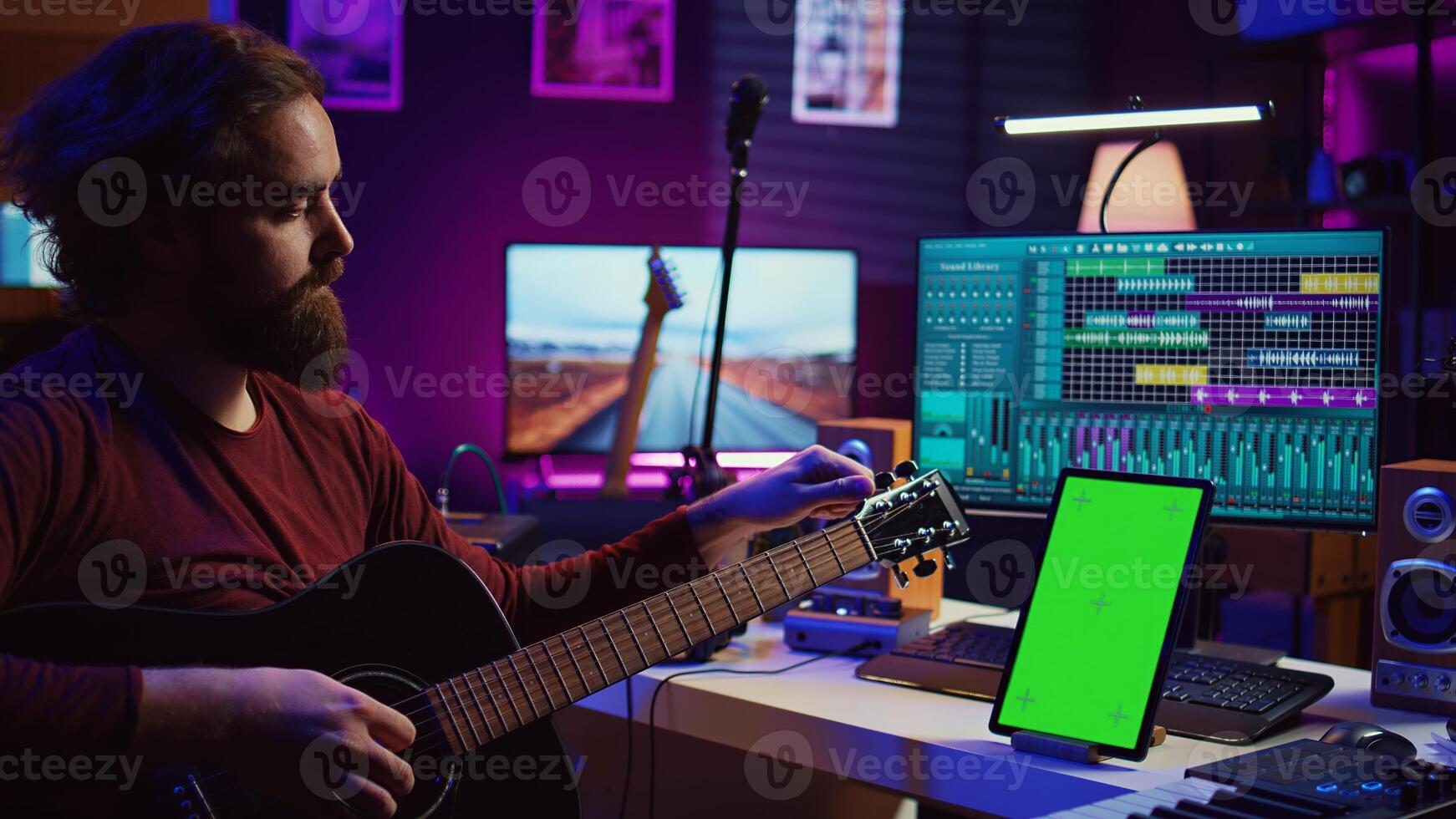Audio engineer practices tuning his acoustic guitar in home studio, watching a tutorial online via tablet with greenscreen. Musician learning to play instrument and create music. Camera B. photo