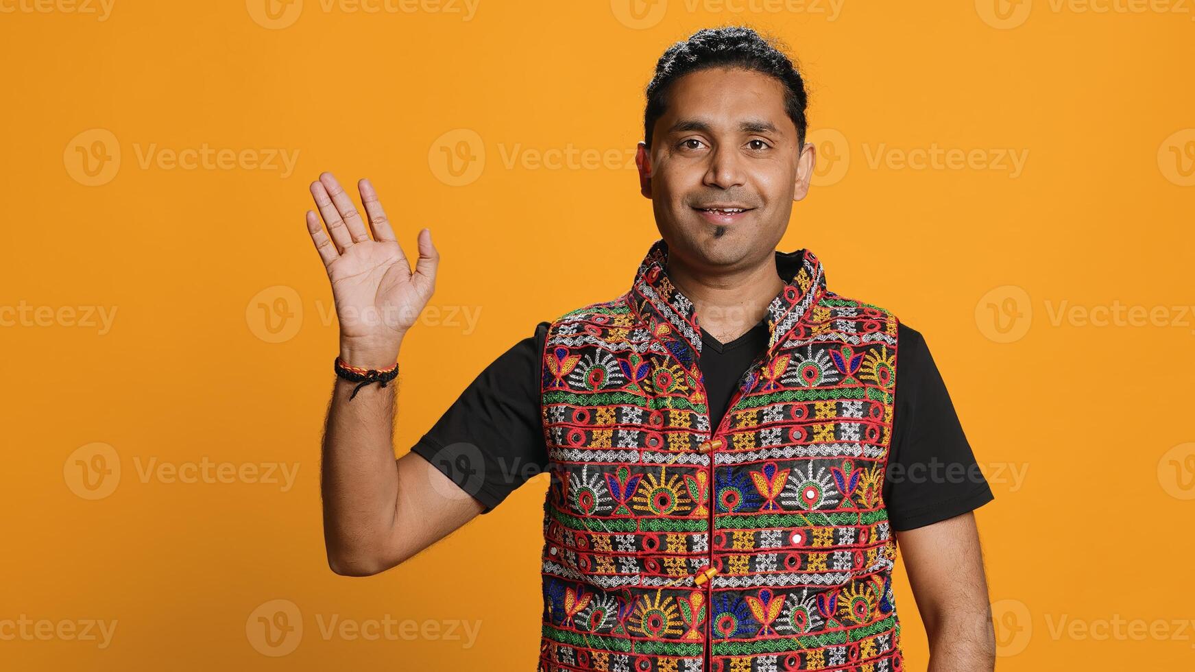 Portrait of jolly man smiling and waving hand, saluting, gesturing, isolated over studio backdrop. Joyous upbeat indian person in traditional attire doing greeting hand gesture, camera A photo
