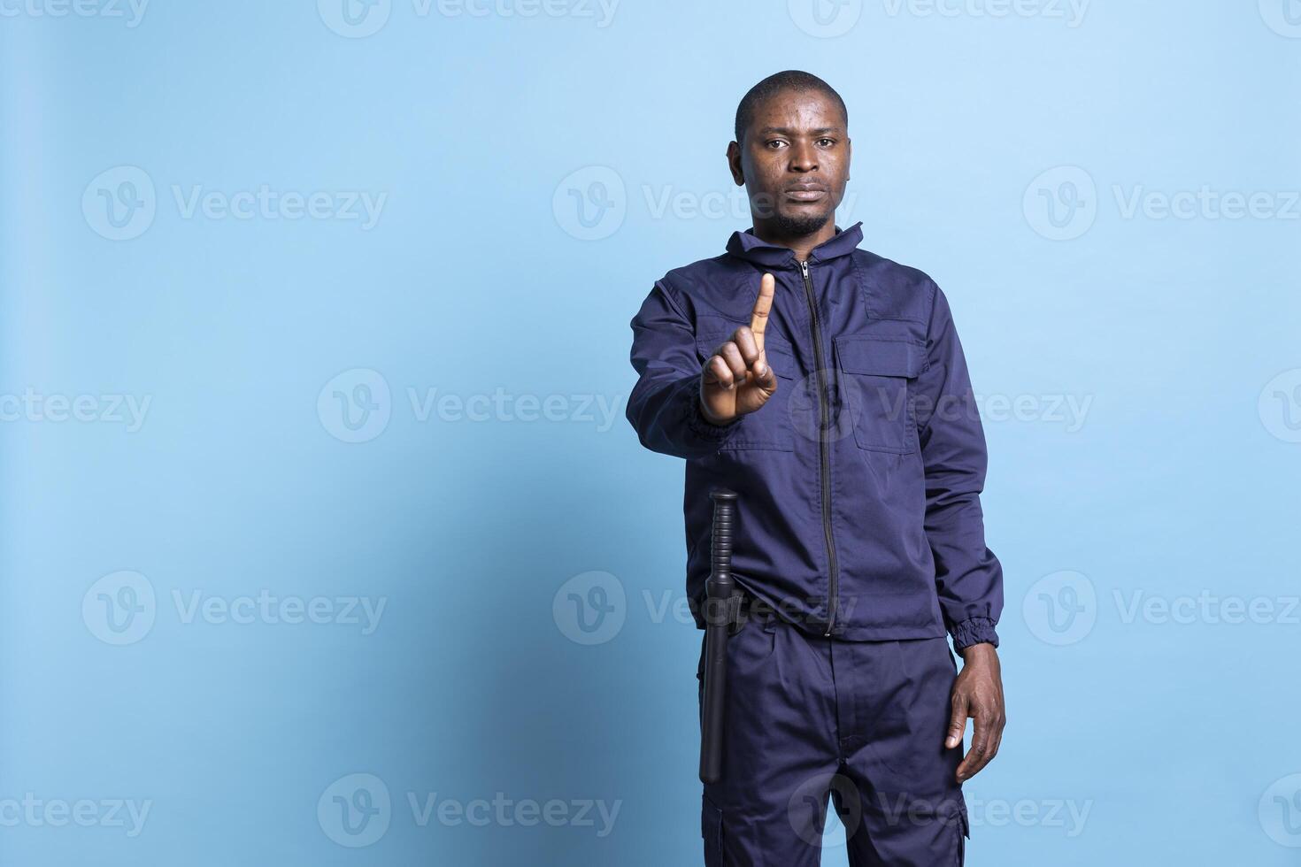 African american security officer raising a finger in front of the camera, showing a restriction symbol against blue background. Man working as a patrol agent presents a stop sign in studio. photo
