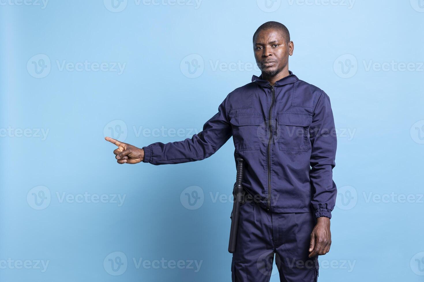 African american man in security agent uniform points to the left or right side, does a commercial advertisement against blue background. Bodyguard indicating a direction with his index finger. photo