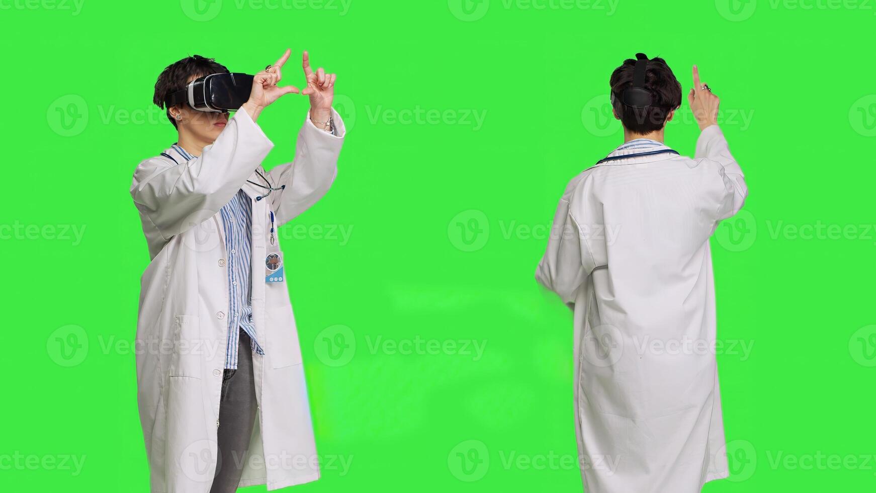 Woman medic works with 3d virtual reality headset in studio, standing against greenscreen backdrop. General practitioner uses vr glasses with interactive vision tool, futuristic exam. Camera B. photo