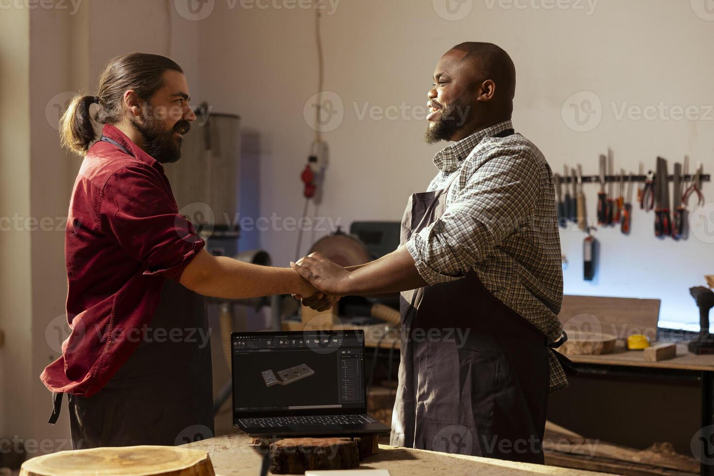 Carpenter happy to meet upbeat client in woodworking shop, discussing commissioned furniture assembling job. Joyous cabinetmaker talking with customer about blueprint on CAD software on laptop photo