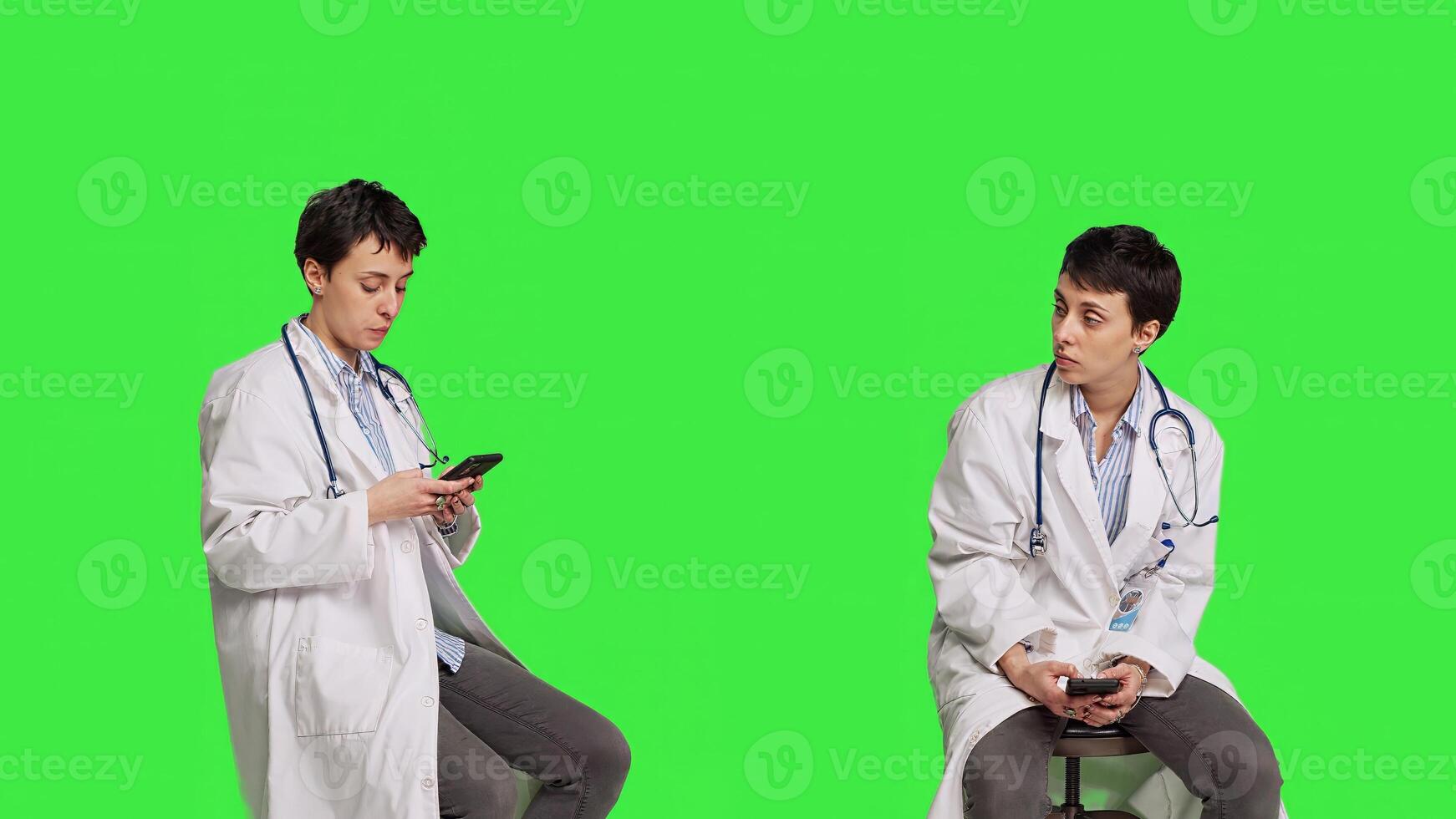 Woman specialist using smartphone social media apps in studio, sitting on a chair against greenscreen backdrop. Physician waiting for someone and browses online webpages, texting. Camera B. photo