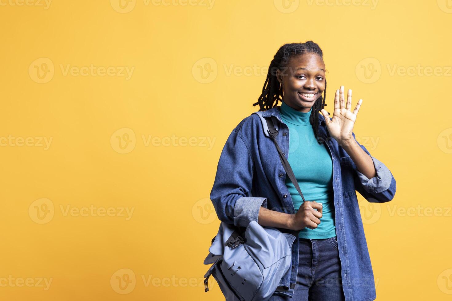 Portrait of smiling girl holding backpack, waving hand, gesturing, isolated over yellow studio background. Joyful african american student with school rucksack doing greeting hand gesture photo