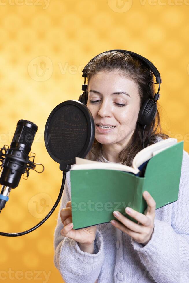 Smiling narrator wearing headphones reading aloud from book into microphone against yellow background. Cheerful person recording audiobook, creating engaging media content for listeners photo