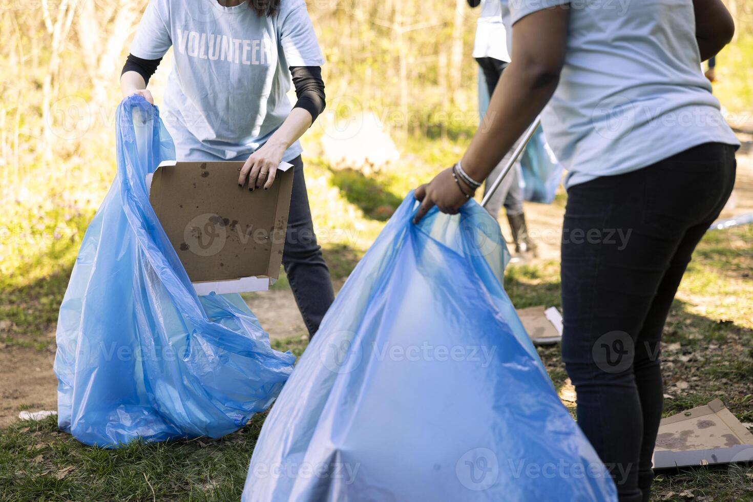 Women volunteers tidying the forest area of junk and plastic waste, collecting garbage with claw and blue disposal bag. Team of activists volunteering to preserve natural environment. photo