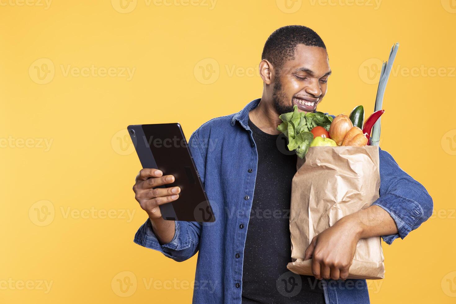 Happy vegan young man browses social media on tablet, shopping for ethically sourced groceries from farmers market. African american person holds natural produce and gadget. photo