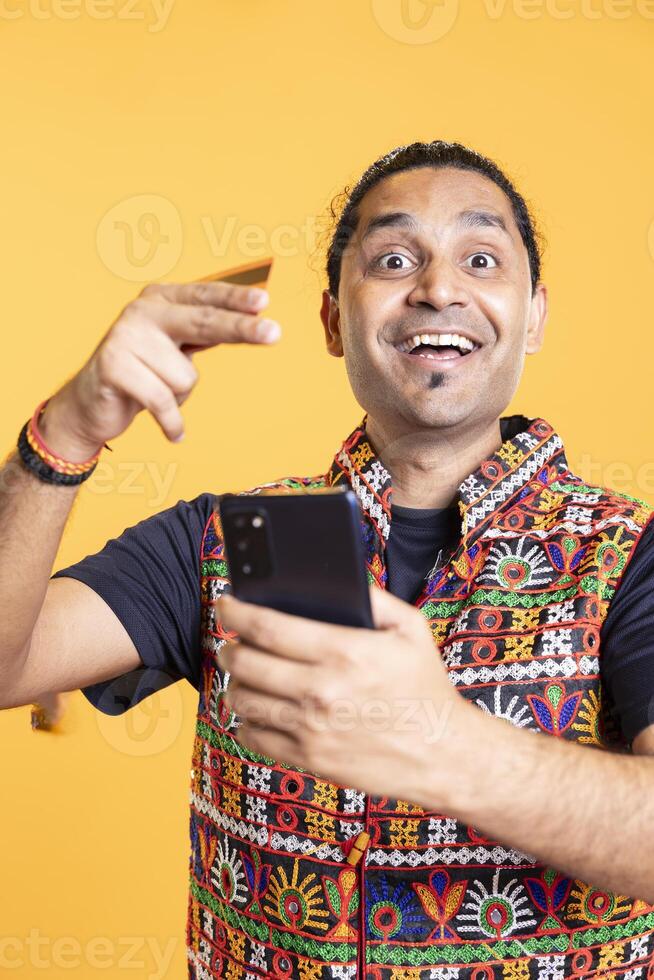 Portrait of jolly man looking at debit card to read banking account details, thrilled to do online purchases, studio background. Joyous man stoked to do internet shopping using cellphone photo